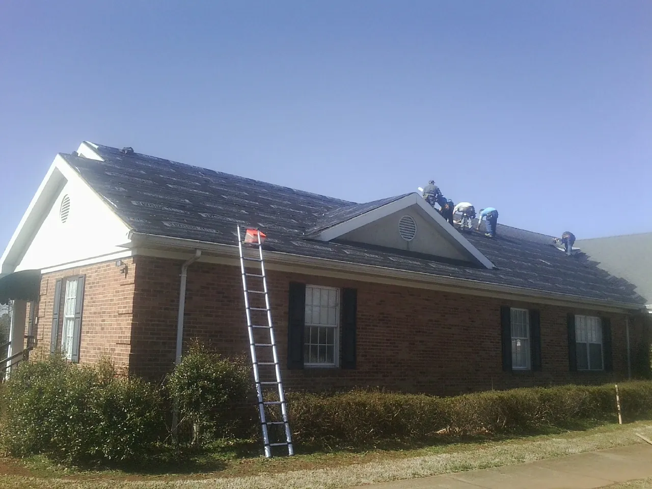 Crew tearing shingles off a brick ranch roof with a ladder set — Emergency Roof Repair work by our crew in Stockport