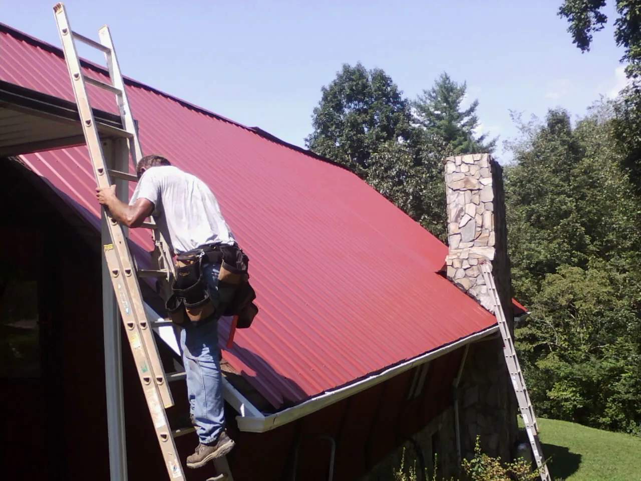 Roofer on a ladder beside a red metal roof and stone chimney — Soffit & Fascia Repair by Roofing Brothers in Stockport